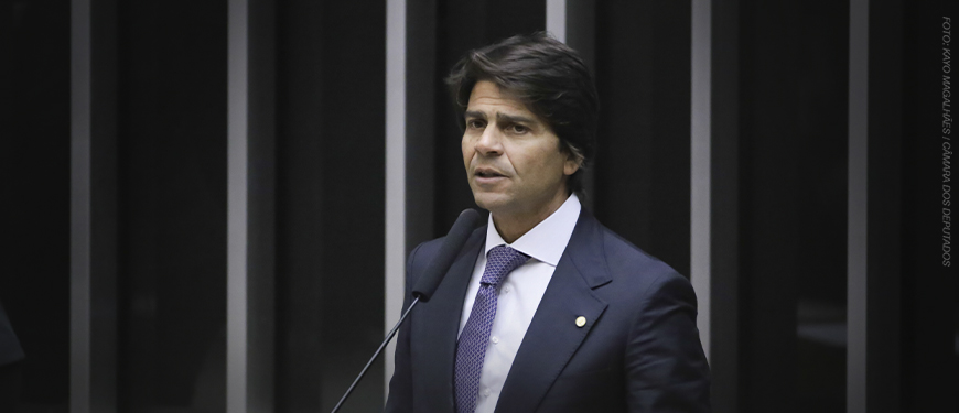 A imagem mostra o Deputado Pedro Paulo de terno falando ao microfone em um ambiente formal.