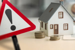 Imagem Acessível: A imagem mostra uma miniatura de casa cercada por moedas empilhadas, representando patrimônio e finanças pessoais. Em primeiro plano, um sinal de alerta vermelho com ponto de exclamação chama atenção para a necessidade de regularização. O texto reforça a orientação: “Saiba se você está na lista para regularizar a Declaração de Bens e Valores”, alertando os servidores sobre a obrigação anual de envio das informações patrimoniais. A arte tem tom informativo e preventivo, destacando a importância da atualização dentro do prazo. Na base da imagem, há a logomarca do SINJUS-MG.