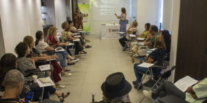 Imagem Acessível: A imagem mostra uma sala com participantes sentados em formato de roda, acompanhando a apresentação de duas facilitadoras à frente, com apoio de projeção e banner do NAP/SINJUS-MG. Em primeiro plano, quatro mulheres sorriem para a foto, segurando os materiais disponibilizados no evento. O texto em destaque informa que o Encontro do NAP abordou o tema da respiração consciente, e anuncia uma mentoria exclusiva, reforçando a iniciativa do NAP voltada à saúde emocional, qualidade de vida e desenvolvimento pessoal dos filiados.