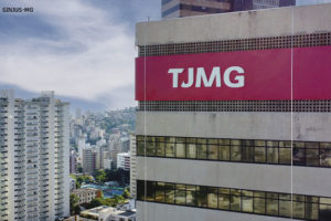 Vista dos últimos andares do edifício sede do Tribunal de Justiça de Minas Gerais, ele tem uma fachada em concreto aparente e vidros espelhados que evidenciam os níveis da construção. No topo, há um letreiro com fundo vermelho onde se lê TJMG. O céu está claro e com algumas nuvens, ao fundo é possível avistar os bairros vizinhos com muitas casas e edifícios.