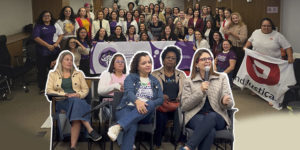 A imagem mostra uma grande reunião de mulheres sindicalistas em um auditório, com bandeiras de diversos sindicatos estendidas no chão. Ao centro, destaca-se a faixa do Encontro de Mulheres Dirigentes Sindicais da FENAJUD. Na parte inferior, em destaque, cinco mulheres do SINJUS-MG sentadas participam ativamente das discussões. O texto informa que o Núcleo das Mulheres do SINJUS foi destaque neste encontro nacional de sindicalistas.