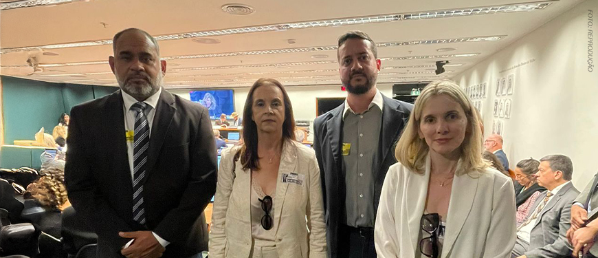 A imagem mostra quatro representantes sindicais posando lado a lado no centro do plenário da Câmara dos Deputados, durante uma audiência. Eles estão vestidos formalmente, com expressões sérias e engajadas. Ao fundo, é possível ver parlamentares e outros participantes sentados, além de uma tela com transmissão da audiência. O destaque da imagem é o texto: “Reforma Administrativa em foco: Fenajud e Sindicatos marcam presença em audiência na Câmara”.