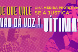 Arte do Agosto Lilás com fundo em tons de rosa e roxo. Em destaque, a pergunta: “De que vale uma medida protetiva se a Justiça não dá voz à vítima?”. Ao fundo, aparecem imagens sobrepostas de uma manifestação de mulheres; à direita, uma pessoa segura um megafone, enquanto, na parte inferior, mulheres participam de um ato público. No canto superior esquerdo, lê-se “Agosto Lilás” e, no canto inferior esquerdo, está o logotipo do Núcleo das Mulheres do SINJUS-MG.