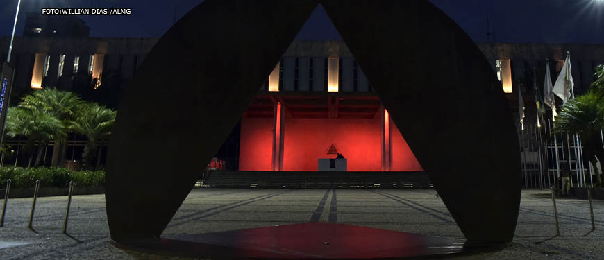 Vista noturna da ALMG (Palácio da Inconfidência), à frente do edifício, há uma escultura onde se vê um triângulo que remete à bandeira do estado de Minas. Pelo triângulo, pode-se ver o edifício iluminado em tons de vermelho. Conteúdo textual: Eleições 2022 - Na ALMG, ala progressista aumentou; no Congresso, cenário é obscuro.