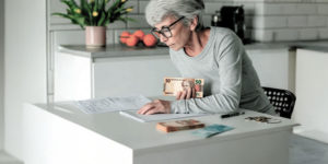 Imagem Acessível: Card com fundo de uma cozinha clara. Uma senhora de cabelos grisalhos e óculos está sentada à mesa, contando notas de dinheiro e analisando documentos.Conteúdo textual: QUITAÇÃO - Em ofício, SINJUS solicita transparência e agilidade no pagamento da URV.