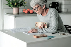 Imagem Acessível: Card com fundo de uma cozinha clara. Uma senhora de cabelos grisalhos e óculos está sentada à mesa, contando notas de dinheiro e analisando documentos.Conteúdo textual: QUITAÇÃO - Em ofício, SINJUS solicita transparência e agilidade no pagamento da URV.