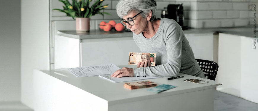 Imagem Acessível: Card com fundo de uma cozinha clara. Uma senhora de cabelos grisalhos e óculos está sentada à mesa, contando notas de dinheiro e analisando documentos.Conteúdo textual: QUITAÇÃO - Em ofício, SINJUS solicita transparência e agilidade no pagamento da URV.