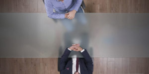 Imagem Acessível: Fotografia aérea de uma mesa de reunião, com duas pessoas sentadas frente a frente — uma com terno escuro e outra com camisa xadrez azul — apoiando as mãos sobre a mesa em posição de diálogo. Conteúdo textual: Grupo de Trabalho - TJMG volta atrás e desmonta avanços na regulamentação da jornada de 8 horas.