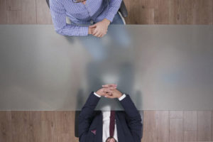Imagem Acessível: Fotografia aérea de uma mesa de reunião, com duas pessoas sentadas frente a frente — uma com terno escuro e outra com camisa xadrez azul — apoiando as mãos sobre a mesa em posição de diálogo. Conteúdo textual: Grupo de Trabalho - TJMG volta atrás e desmonta avanços na regulamentação da jornada de 8 horas.