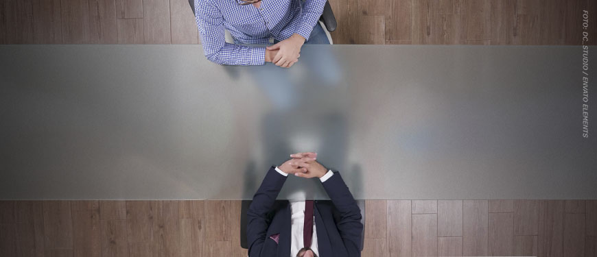 Imagem Acessível: Fotografia aérea de uma mesa de reunião, com duas pessoas sentadas frente a frente — uma com terno escuro e outra com camisa xadrez azul — apoiando as mãos sobre a mesa em posição de diálogo. Conteúdo textual: Grupo de Trabalho - TJMG volta atrás e desmonta avanços na regulamentação da jornada de 8 horas.
