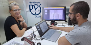 Duas pessoas trabalham lado a lado em um escritório. Uma mulher de óculos de proteção transparente sorri para a câmera, enquanto um homem de barba utiliza um notebook conectado a um monitor externo que exibe documentos digitalizados. Na parede aparece o logo ‘Plantão da PV 2024’. Sobre a mesa há papéis e equipamentos de trabalho.