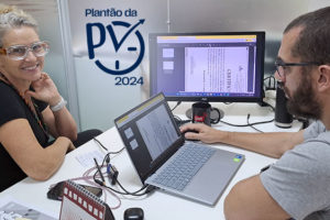 Duas pessoas trabalham lado a lado em um escritório. Uma mulher de óculos de proteção transparente sorri para a câmera, enquanto um homem de barba utiliza um notebook conectado a um monitor externo que exibe documentos digitalizados. Na parede aparece o logo ‘Plantão da PV 2024’. Sobre a mesa há papéis e equipamentos de trabalho.