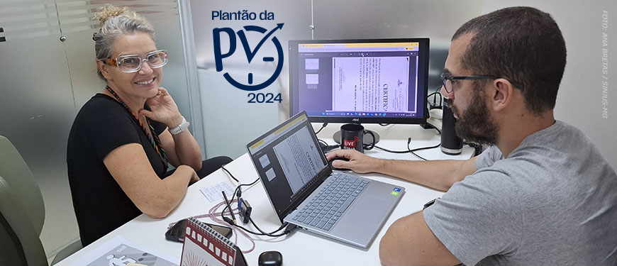 Duas pessoas trabalham lado a lado em um escritório. Uma mulher de óculos de proteção transparente sorri para a câmera, enquanto um homem de barba utiliza um notebook conectado a um monitor externo que exibe documentos digitalizados. Na parede aparece o logo ‘Plantão da PV 2024’. Sobre a mesa há papéis e equipamentos de trabalho.
