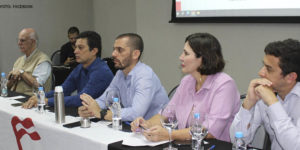 Imagem fotografia de uma sala com pessoas sentadas em cadeiras e a frente uma mesa, sob a mesma, uma bandeira da Fenajud se vê ao fundo um telão com o título "Coletivo Jurídico".