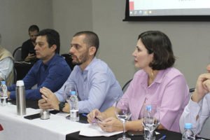 Imagem fotografia de uma sala com pessoas sentadas em cadeiras e a frente uma mesa, sob a mesma, uma bandeira da Fenajud se vê ao fundo um telão com o título "Coletivo Jurídico".