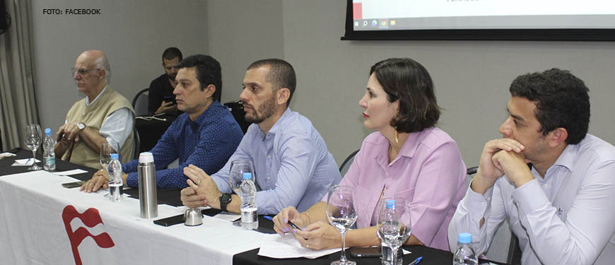 Imagem fotografia de uma sala com pessoas sentadas em cadeiras e a frente uma mesa, sob a mesma, uma bandeira da Fenajud se vê ao fundo um telão com o título "Coletivo Jurídico".