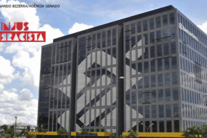 Fotografia diurna da fachada do Banco do Brasil com sede em Brasília, na lateral da imagem se ve o logo Sinjus antirracista na cor vermelha.