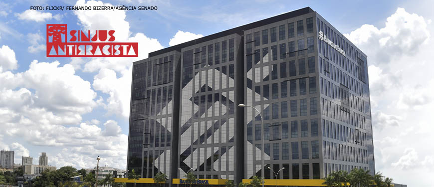 Fotografia diurna da fachada do Banco do Brasil com sede em Brasília, na lateral da imagem se ve o logo Sinjus antirracista na cor vermelha.