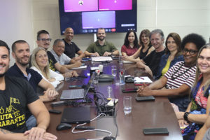 Diretores da diretoria colegiada do SINJUS reunidos em uma sala de reuniões, sentados ao redor de uma mesa retangular. O grupo, composto por homens e mulheres, sorri para a câmera em um ambiente institucional, com notebooks, celulares, copos de água e equipamentos de reunião sobre a mesa. Ao fundo, há uma tela com videoconferência exibindo participantes remotos.