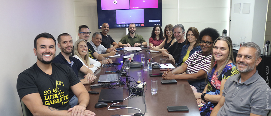 Diretores da diretoria colegiada do SINJUS reunidos em uma sala de reuniões, sentados ao redor de uma mesa retangular. O grupo, composto por homens e mulheres, sorri para a câmera em um ambiente institucional, com notebooks, celulares, copos de água e equipamentos de reunião sobre a mesa. Ao fundo, há uma tela com videoconferência exibindo participantes remotos.