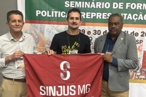 Imagem acessível: A imagem mostra o representante do SINJUS-MG segurando a bandeira do sindicato durante o Seminário de Formação Política e Representação de Classe, promovido pela CSPB, em ambiente interno com banner do evento ao fundo. No topo lê-se “Movimento nacional”. Na parte inferior o texto informa que o SINJUS participou do seminário e reforçou o debate sobre a atuação política dos sindicatos.