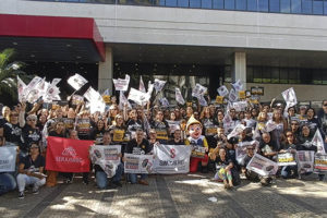 A imagem mostra um grande número de servidoras e servidores, além de dirigentes do SINJUS e do SERJUSMIG, em frente à sede do TJMG com faixas e cartazes em protesto contra o descumprimento de acordos e direitos.