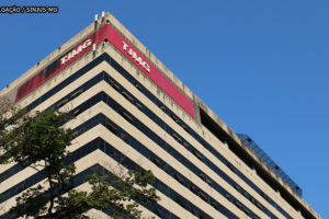 Fachada da frente do edifício sede do Tribunal de Justiça de Minas Gerais (TJMG). A perspectiva da imagem é de baixo para cima, na qual se vê a marte superior do prédio e no último andar um letreiro branco sobre fundo vermelho com a sigla TJMG. Ao fundo do edifício o céu está azul e sem nuvens.