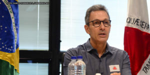 Governador de Minas Gerais, Romeu Zema (homem de pele clara, cabelos grisalhos curtos e usando óculos preto e camisa cinza) sentado entre as bandeiras do Brasil e de Minas Gerais.