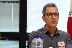 Governador de Minas Gerais, Romeu Zema (homem de pele clara, cabelos grisalhos curtos e usando óculos preto e camisa cinza) sentado entre as bandeiras do Brasil e de Minas Gerais.