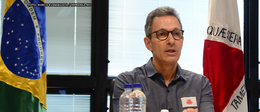 Governador de Minas Gerais, Romeu Zema (homem de pele clara, cabelos grisalhos curtos e usando óculos preto e camisa cinza) sentado entre as bandeiras do Brasil e de Minas Gerais.