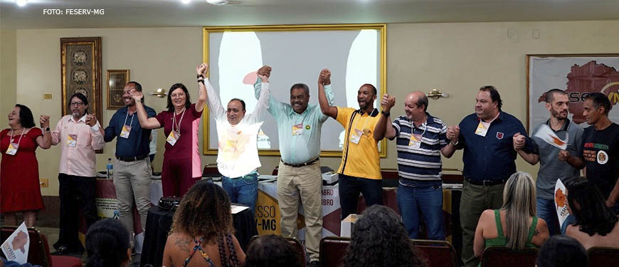 Representantes de entidades sindicais de servidores públicos municipais reunidos perante uma plateia, eles se dão as mãos e estão com os braços levantados; representando o SINJUS-MG estão o coordenador-geral, Alexandre Pires e o diretor de Assuntos Sociais, Culturais e de Saúde, Jonas Araújo.