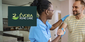 Imagem Acessível: A imagem mostra uma enfermeira atendendo um homem em sua casa, o auxiliando enquanto ele se levanta do sofá, ambos sorrindo. Sobre a imagem, aparece o texto: ‘Tenha atendimento médico domiciliar com o novo convênio do SINJUS’. No canto inferior esquerdo está o logo da empresa Évora Home Care.