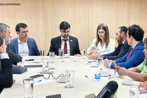 Imagem fotográfica de uma sala de reunião com uma mesa onde os participantes se encontram sentados com fisionomia de debater o assunto, acima da mesa se vê papéis e um microfone. Estão na foto deputados estaduais mineiros e representantes do Ministério da Fazenda.