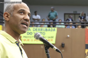 Imagem Acessível: Ao fundo há um ambiente com faixas e um mezanino onde pessoas assistem a um pronunciamento. Em primeiro plano está Wagner Ferreira falando ao microfone, ele é um homem negro, calvo com cabelos grisalhos, tem a barba feita e usa uma camisa pólo amarela.