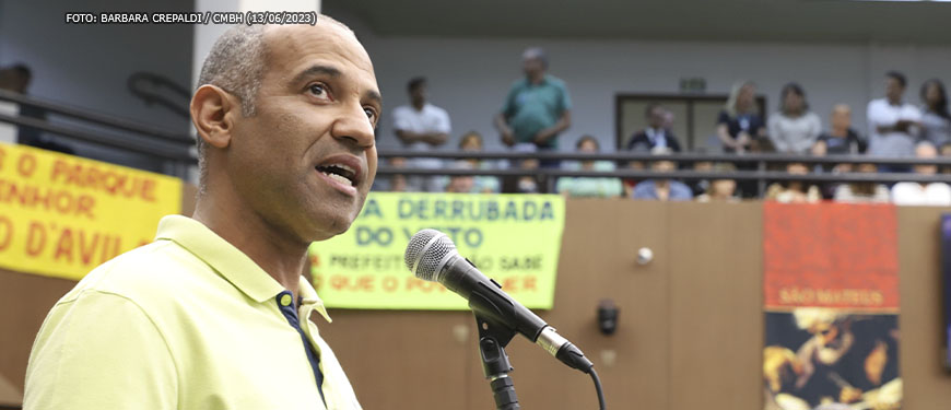 Imagem Acessível: Ao fundo há um ambiente com faixas e um mezanino onde pessoas assistem a um pronunciamento. Em primeiro plano está Wagner Ferreira falando ao microfone, ele é um homem negro, calvo com cabelos grisalhos, tem a barba feita e usa uma camisa pólo amarela.