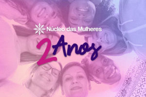 Foto com diversas mulheres abraçadas formando um círculo em tons de rosa e azul de 2º plano, no primeiro plano temos o conteúdo textual " Núcleo das mulheres" " 2 anos"