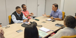 A imagem mostra uma reunião em andamento, com sete pessoas sentadas ao redor de uma mesa retangular em uma sala de reunião. Um dos representantes do SINJUS, em destaque à esquerda, fala enquanto os demais acompanham com atenção.
