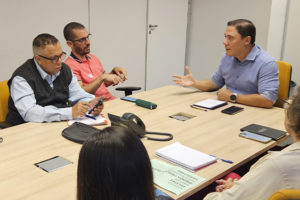 A imagem mostra uma reunião em andamento, com sete pessoas sentadas ao redor de uma mesa retangular em uma sala de reunião. Um dos representantes do SINJUS, em destaque à esquerda, fala enquanto os demais acompanham com atenção.