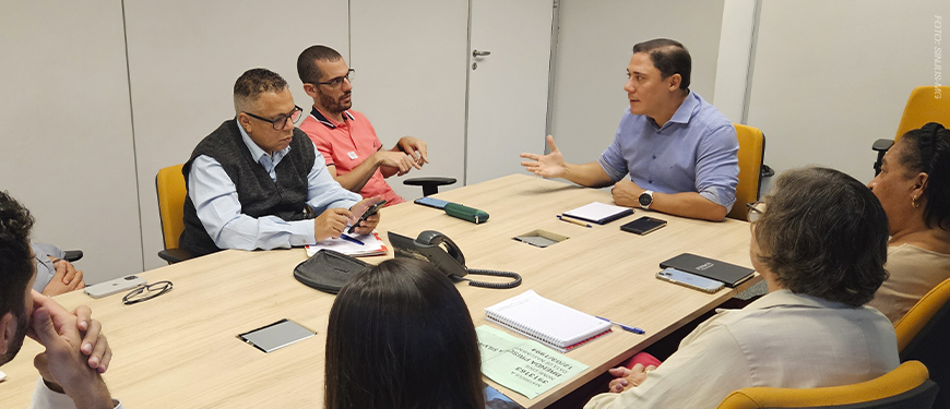 A imagem mostra uma reunião em andamento, com sete pessoas sentadas ao redor de uma mesa retangular em uma sala de reunião. Um dos representantes do SINJUS, em destaque à esquerda, fala enquanto os demais acompanham com atenção.