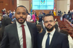 Na imagem há 2 homens em um auditório ambos com trajes sociais; o homem que está do lado esquerdo é negro de cabelos castanhos curtos, ele usa barba e bigode (Valeriano Santos Filho); o outro é branco, tem cabelos castanhos curtos e também usa barba e bigode (Felipe Rodrigues). Ao fundo há um painel de led com um brasão onde se lê " Estado do Rio Grande do Sul - Poder Judiciário - Tribunal de Justiça.