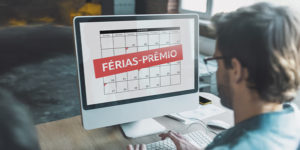 A imagem mostra um homem visto de costas utilizando um computador. Na tela, há um calendário com o texto “FÉRIAS-PRÊMIO” em destaque sobre uma faixa vermelha.