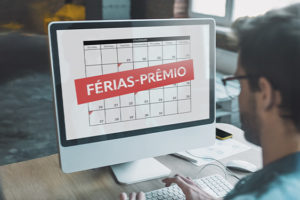 A imagem mostra um homem visto de costas utilizando um computador. Na tela, há um calendário com o texto “FÉRIAS-PRÊMIO” em destaque sobre uma faixa vermelha.