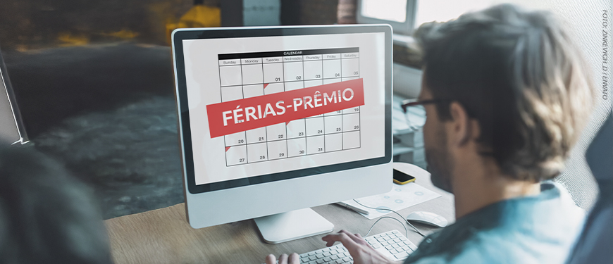 A imagem mostra um homem visto de costas utilizando um computador. Na tela, há um calendário com o texto “FÉRIAS-PRÊMIO” em destaque sobre uma faixa vermelha.