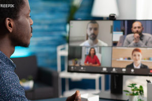 Homem negro com camisa azul de frente para uma tela de computador na qual se vê o rosto desfocado de quatro outras pessoas (dois homens e duas mulheres) que participam de uma reunião por videoconferência.