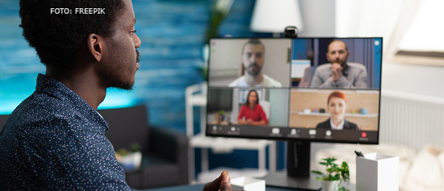 Homem negro com camisa azul de frente para uma tela de computador na qual se vê o rosto desfocado de quatro outras pessoas (dois homens e duas mulheres) que participam de uma reunião por videoconferência.