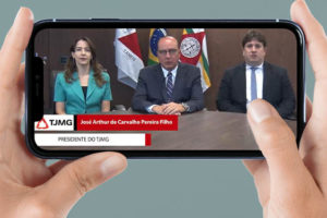 Uma pessoa branca segura um smartphone na horizontal em suas mãos. Na tela há a aplicação digital do vídeo com o anúncio do presidente do TJMG, desembargador José Arthur Filho, homem branco, calvo e de olhos claros, vestido socialmente. Ele está sentado em uma mesa, ao centro, junto de outros membros da direção do Tribunal e olha para a câmera enquanto faz o anúncio aos sindicatos. Conteúdo textual: ATENDENDO AOS SINDICATOS, TRIBUNAL ANUNCIA DATA-BASE 2022 E OUTRAS MEDIDAS