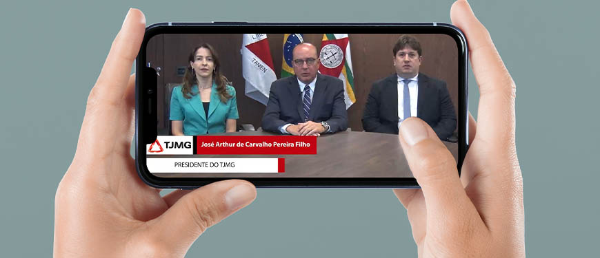 Uma pessoa branca segura um smartphone na horizontal em suas mãos. Na tela há a aplicação digital do vídeo com o anúncio do presidente do TJMG, desembargador José Arthur Filho, homem branco, calvo e de olhos claros, vestido socialmente. Ele está sentado em uma mesa, ao centro, junto de outros membros da direção do Tribunal e olha para a câmera enquanto faz o anúncio aos sindicatos. Conteúdo textual: ATENDENDO AOS SINDICATOS, TRIBUNAL ANUNCIA DATA-BASE 2022 E OUTRAS MEDIDAS