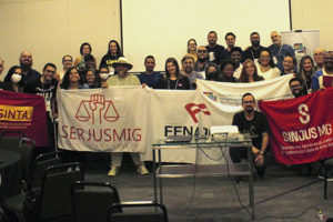 Grupo de mulheres e homens posam para foto em um auditório, elas estão com bandeiras do Encontro de Comunicadores da Fenajud, da Fenajud, do SINJUS, do Serjusmig e do Sintaj.
