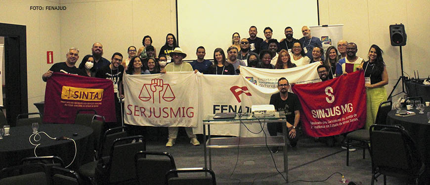 Grupo de mulheres e homens posam para foto em um auditório, elas estão com bandeiras do Encontro de Comunicadores da Fenajud, da Fenajud, do SINJUS, do Serjusmig e do Sintaj.