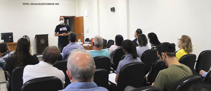 Foto de um auditório com diversos servidores assentados ouvindo o Wagner Ferreira, diretor de Assuntos Jurídicos SINJUS-MG (homem negro, com cabelo bem curto, usa camisa preta e máscara).